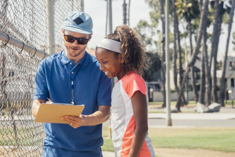 Teacher with clipboard talking to teenage schoolgirl soccer player on school sports field
